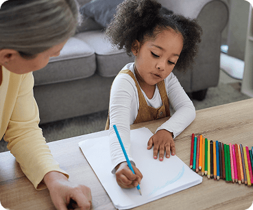 Young girl sits at a table drawing in a notebook with a blue pencil while an adult watches; a row of colored pencils lies on the table nearby.