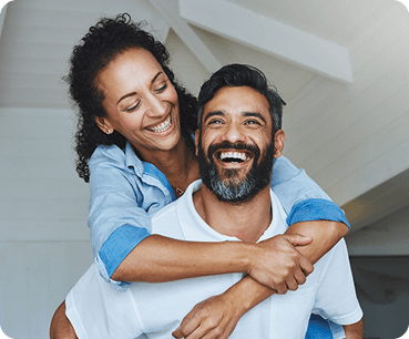 Joyful couple smiling at the camera as the woman hugs the man from behind in a bright home setting